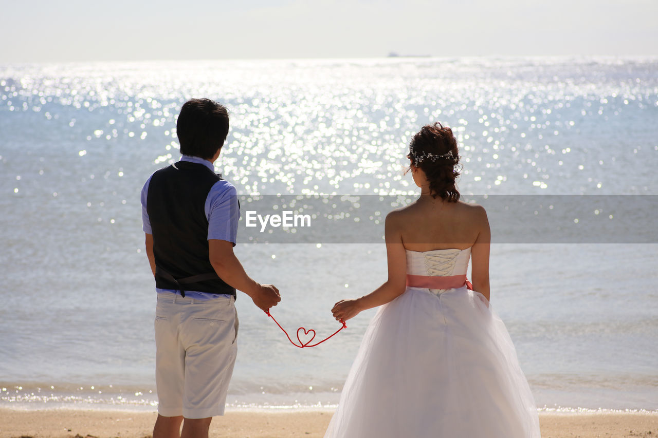 Rear view of couple holding heart shape made with rope while standing at beach