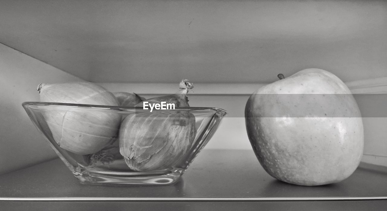 Close-up of onions in bowl and apple on shelf