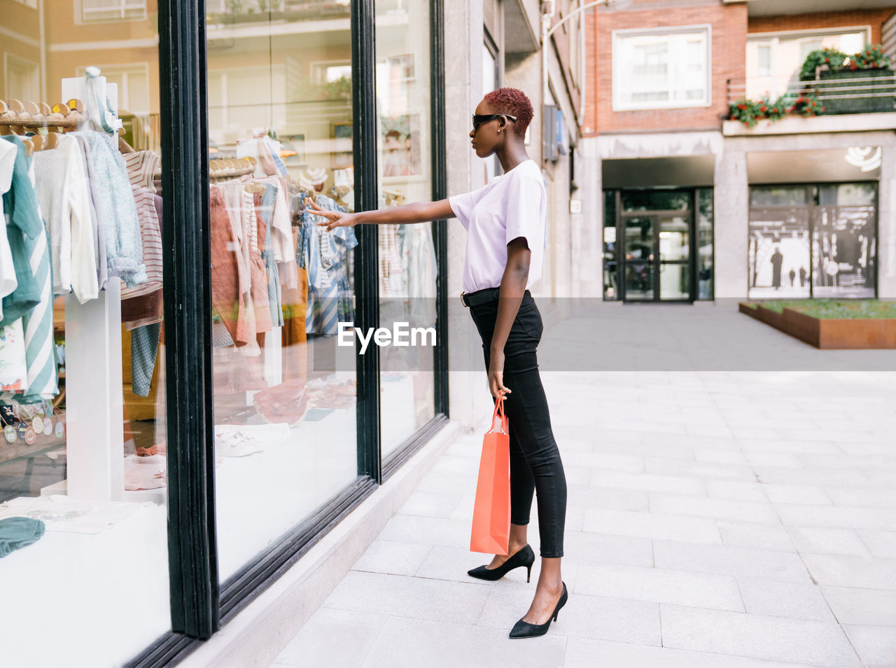 Full body side view of young slim african american female shopper in stylish outfit with shopping bag in hand standing near window of modern clothing store and looking at new collection