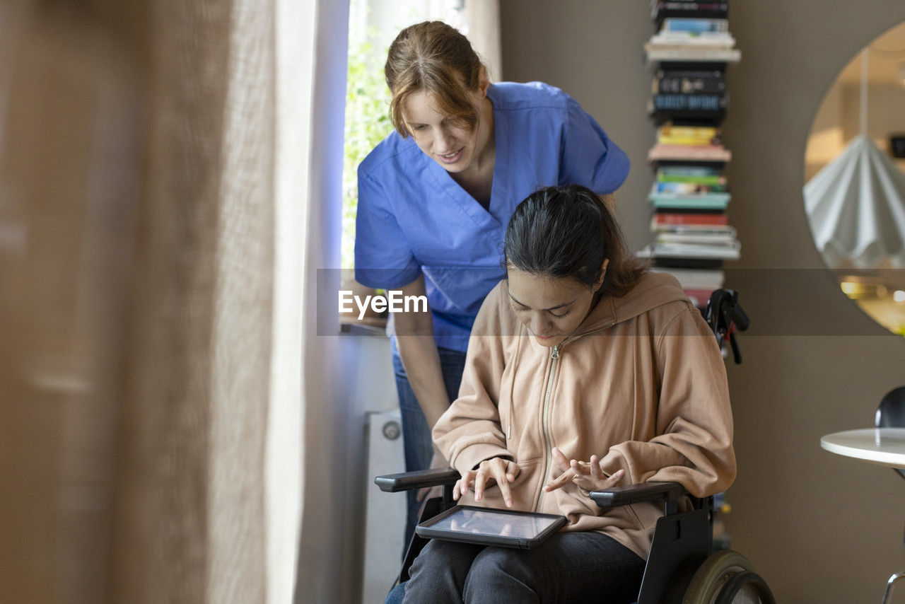 Female caregiver assisting woman using tablet pc while sitting on wheelchair at home