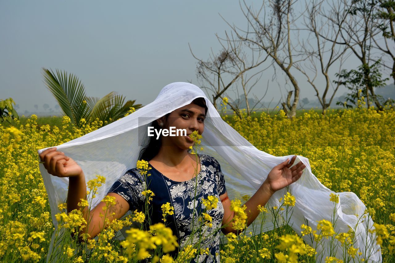 Portrait of smiling teenage girl with scarf standing amidst flowers against sky