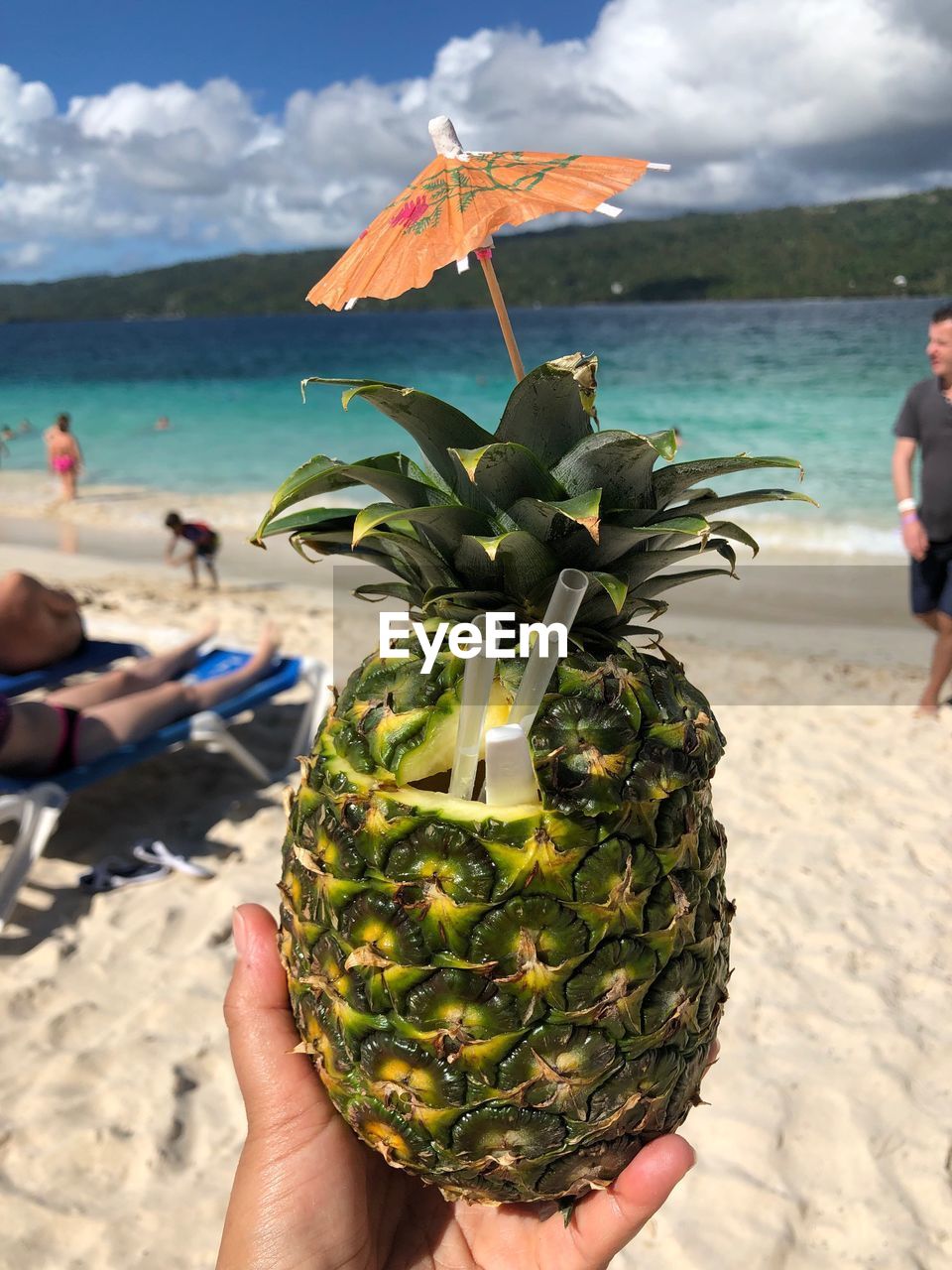 Cropped hand holding pineapple at beach against sky