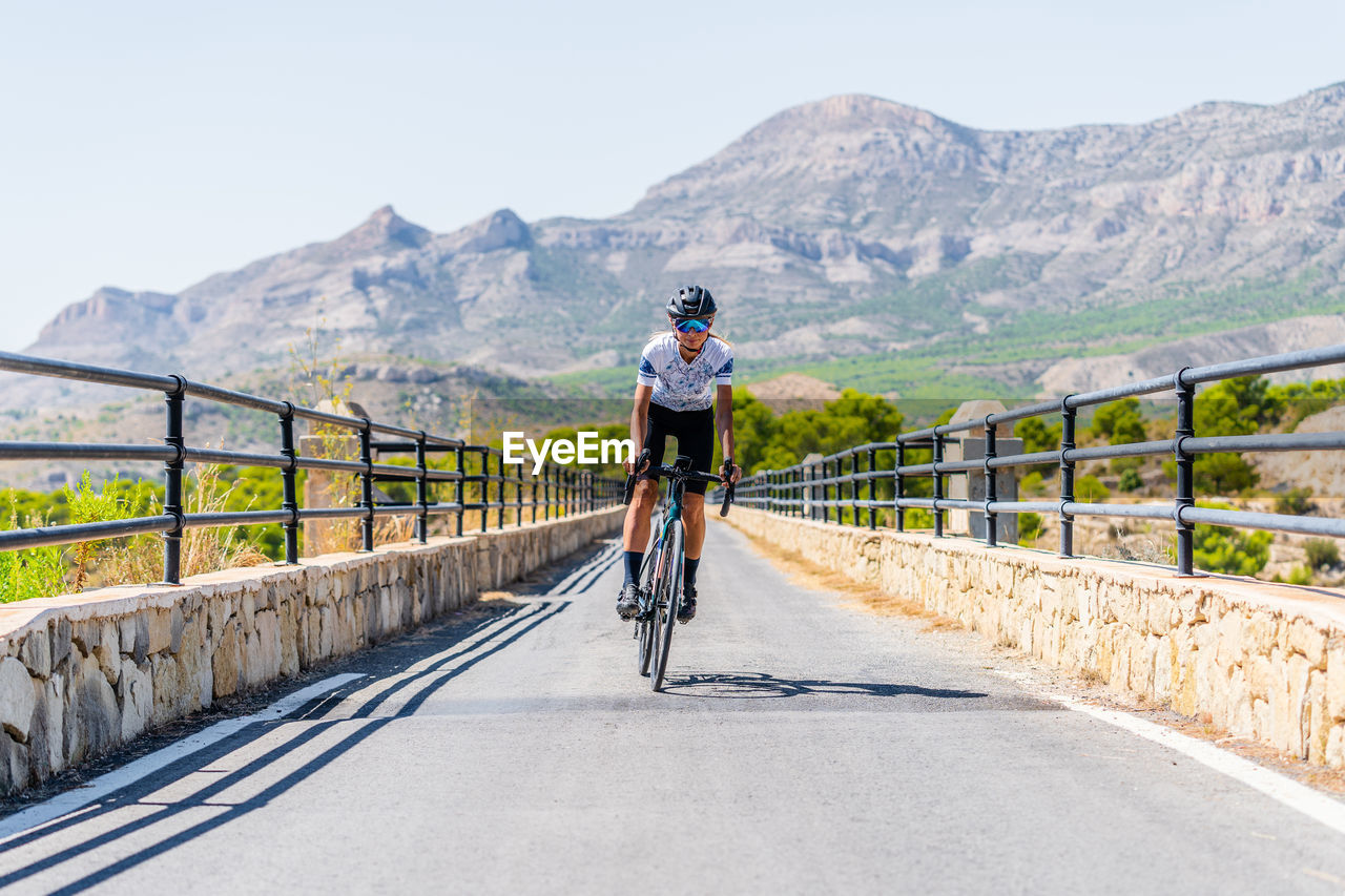 Full length of active female bicyclist in helmet and sportswear riding bike on asphalt road near mountain ridge in summer day