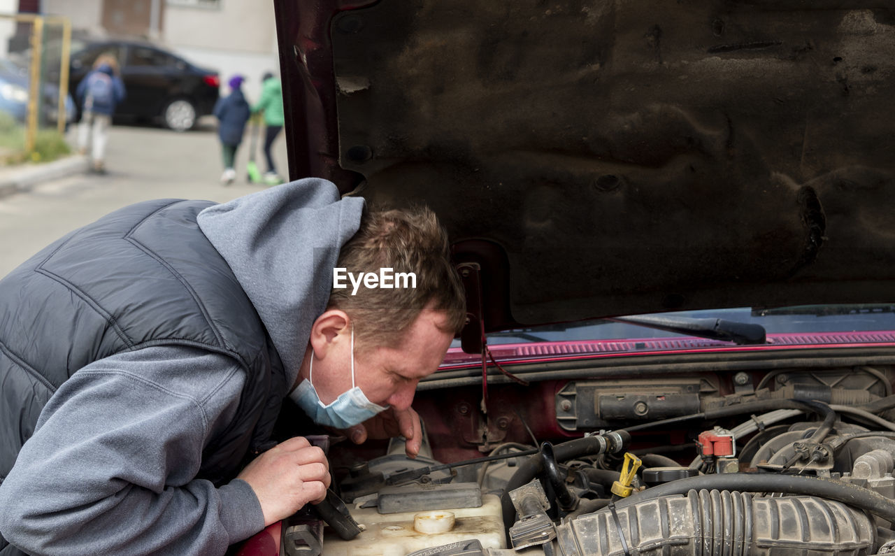 Mechanic in a protective medical mask checks a car engine during a covid-19 coronavirus pandemic