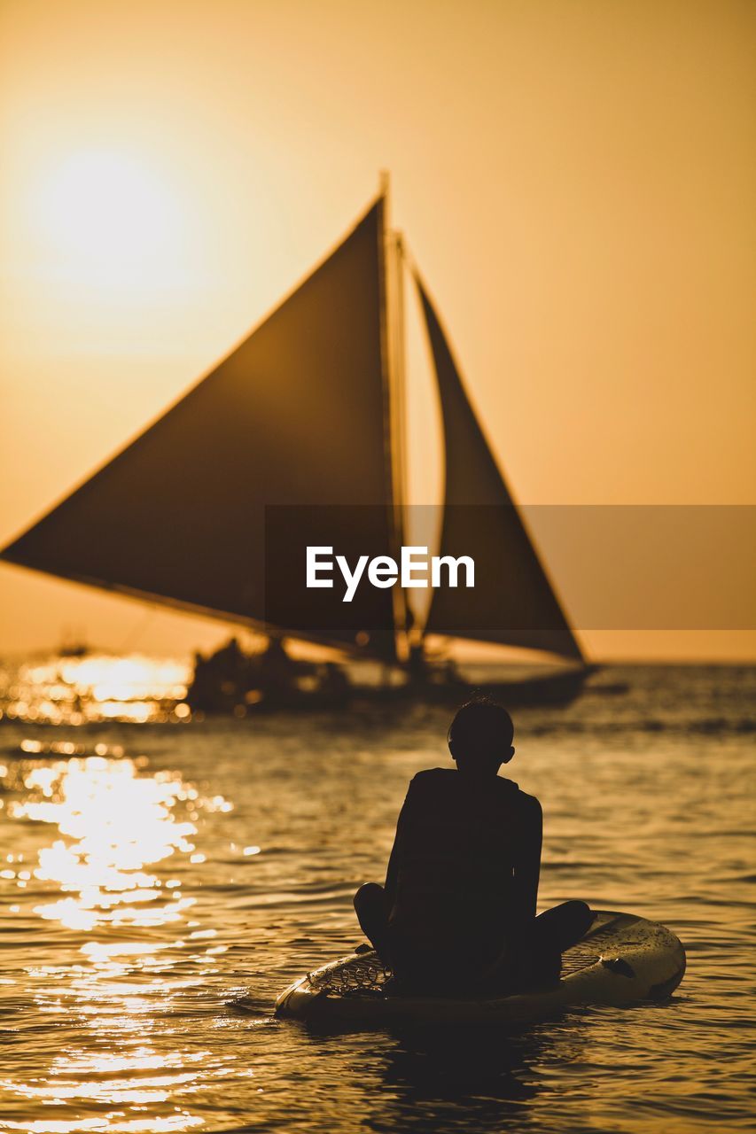 Silhouette boy paddleboarding in sea against sky during sunset