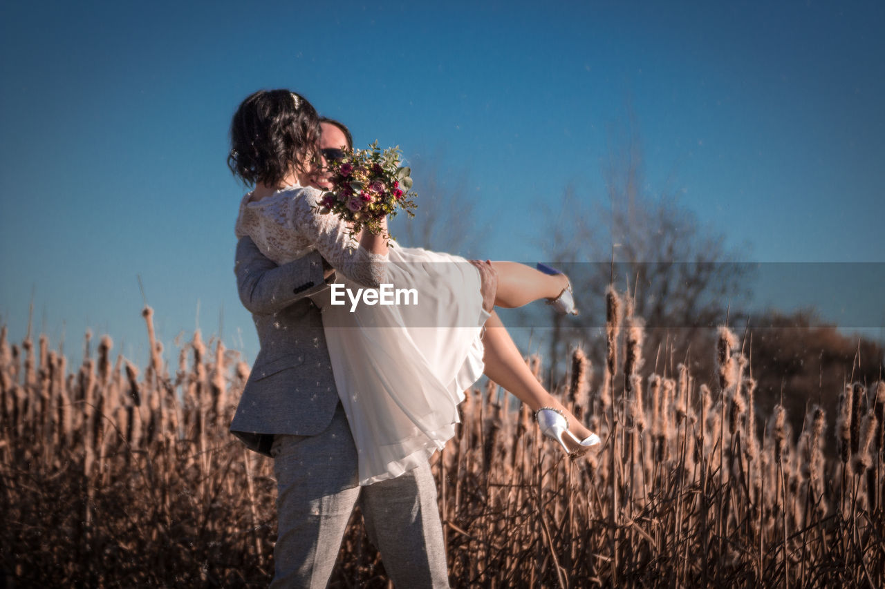 Groom carrying bride holding bouquet on land against sky