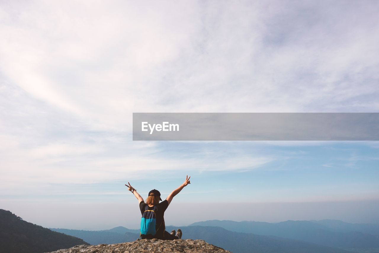 Woman gesturing peace sign while sitting on mountain against sky