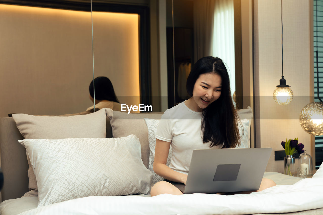 young woman using laptop while sitting on bed at home