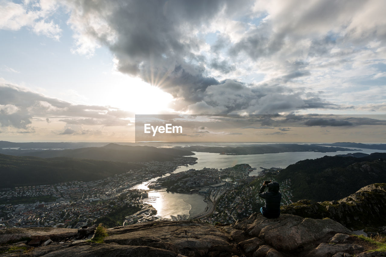 Rear view of man photographing while sitting on rock against sky at sunset