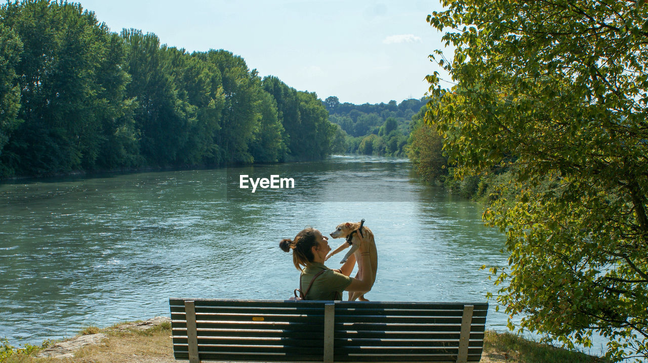 Side view of young woman carrying puppy while sitting on bench by lake against trees