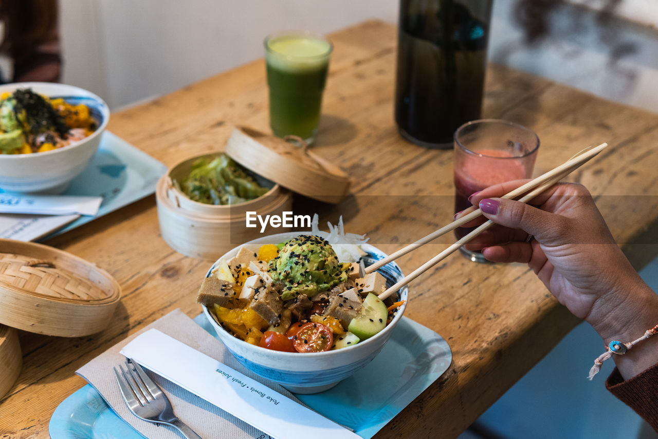 From above of crop anonymous female eating delicious poke dish with chopsticks while sitting at wooden table in restaurant