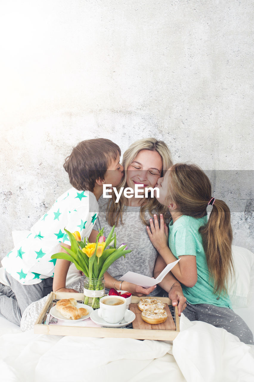 Children kissing mother while having breakfast on bed