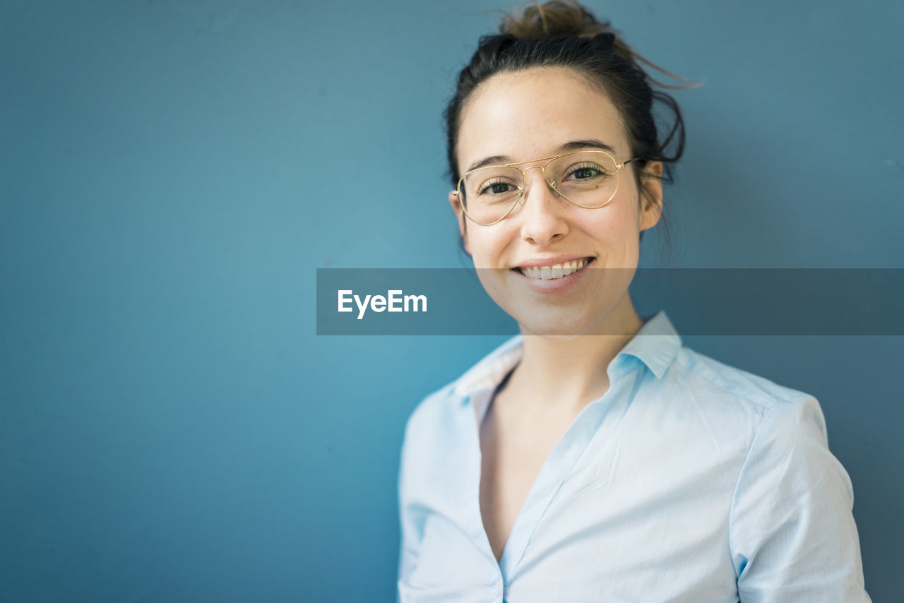 Portrait of smiling young woman wearing glasses in front of blue wall