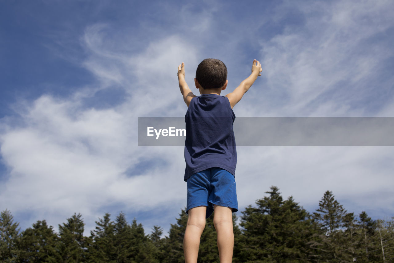 Rear view of boy with arms raised standing against sky