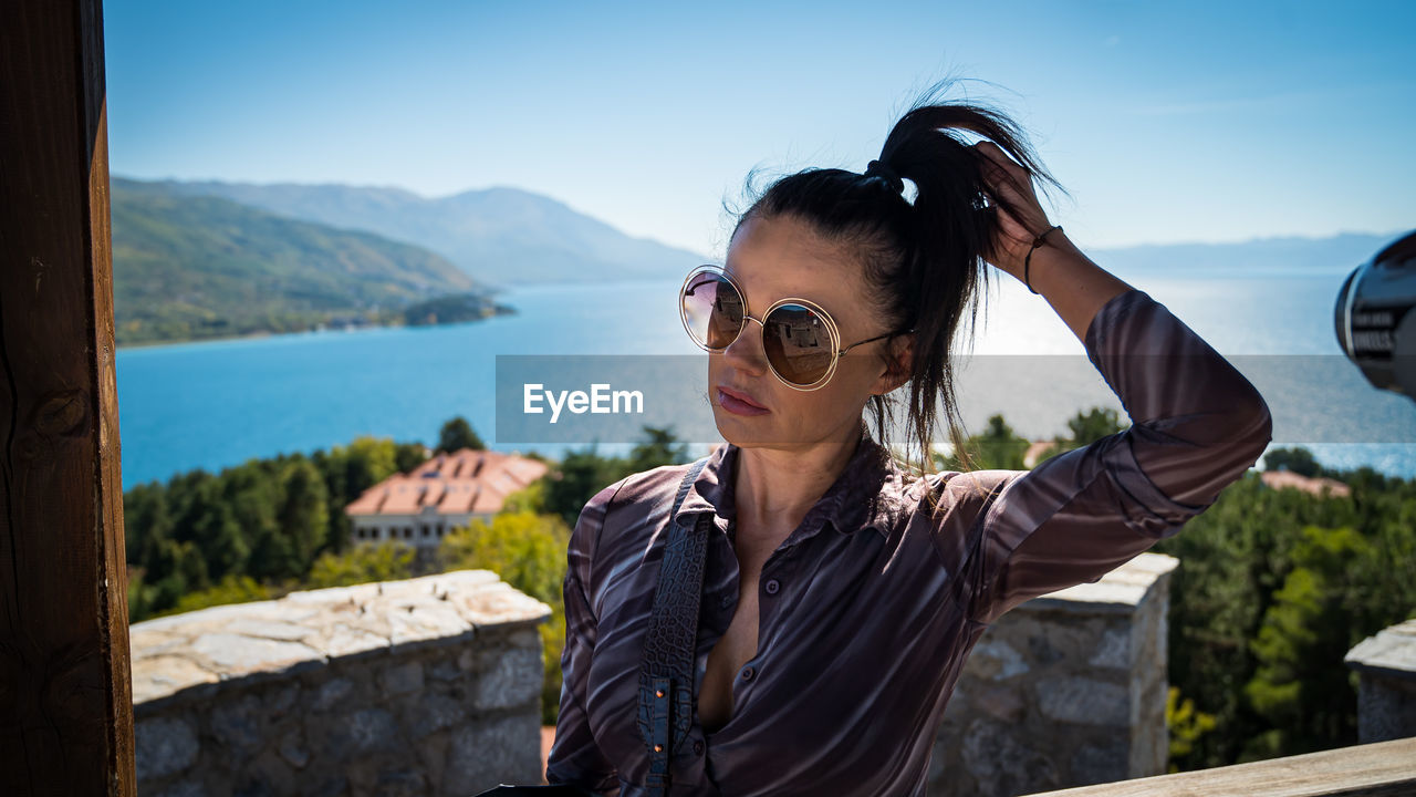 sunglasses, one person, glasses, adult, fashion, nature, sky, water, vacation, mountain, day, travel, women, leisure activity, portrait, young adult, trip, travel destinations, casual clothing, outdoors, lifestyles, holiday, sunlight, goggles, blue, tourism, sea, standing, waist up, beauty in nature, architecture, scenics - nature, mountain range, headshot, relaxation, tourist, looking, land, front view, hairstyle, clothing, tranquility, landscape, female, sunny, focus on foreground