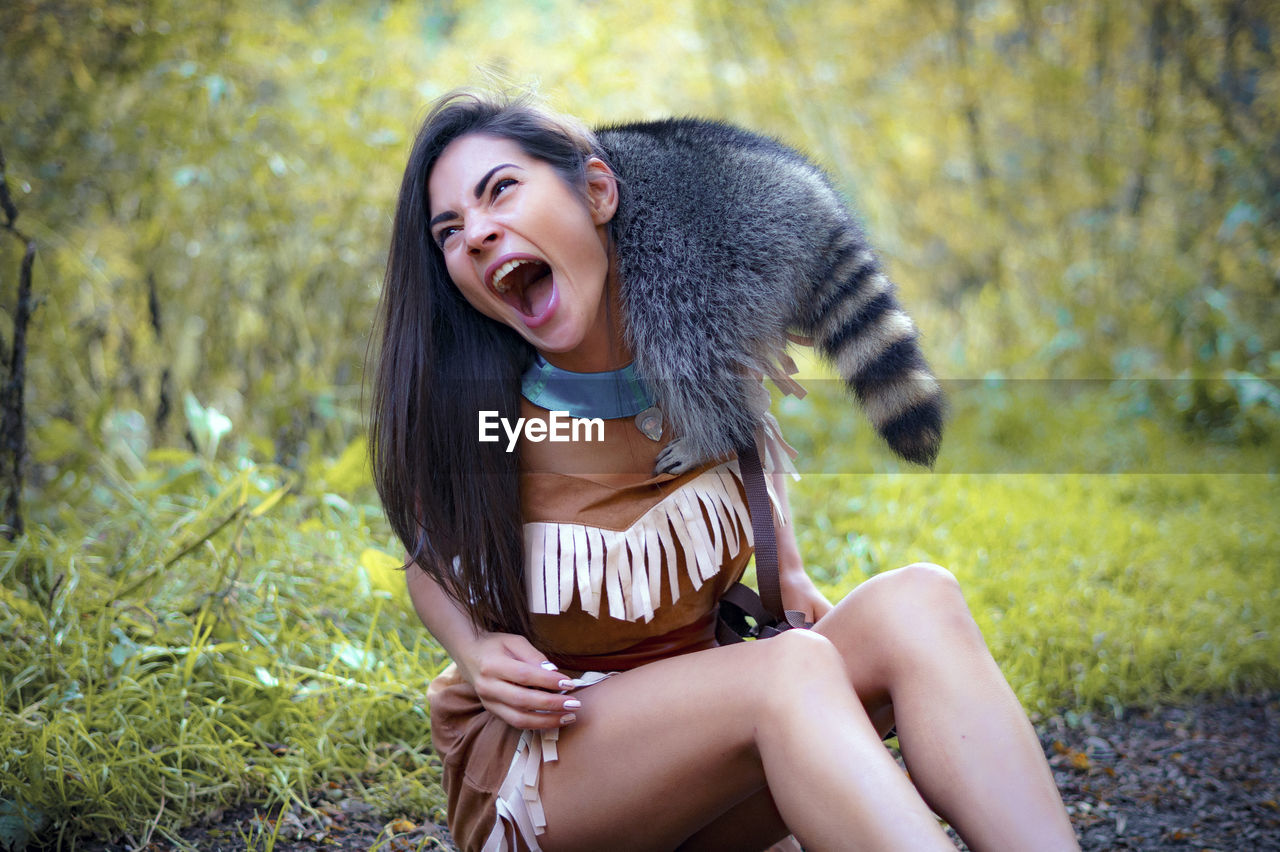 Young woman in traditional clothing playing with raccoon in forest