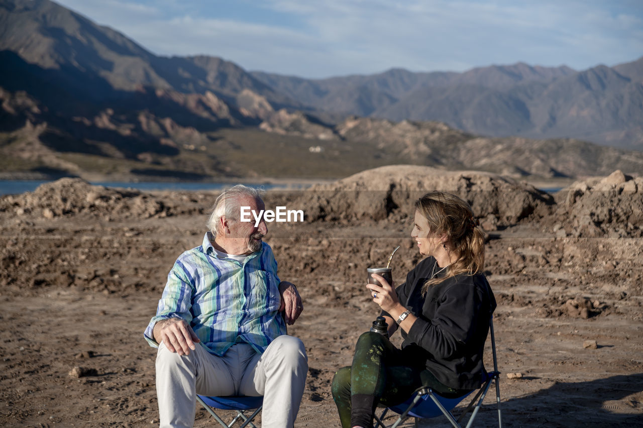 Woman and her grandfather talking while sitting on camping chairs outdoors in nature.