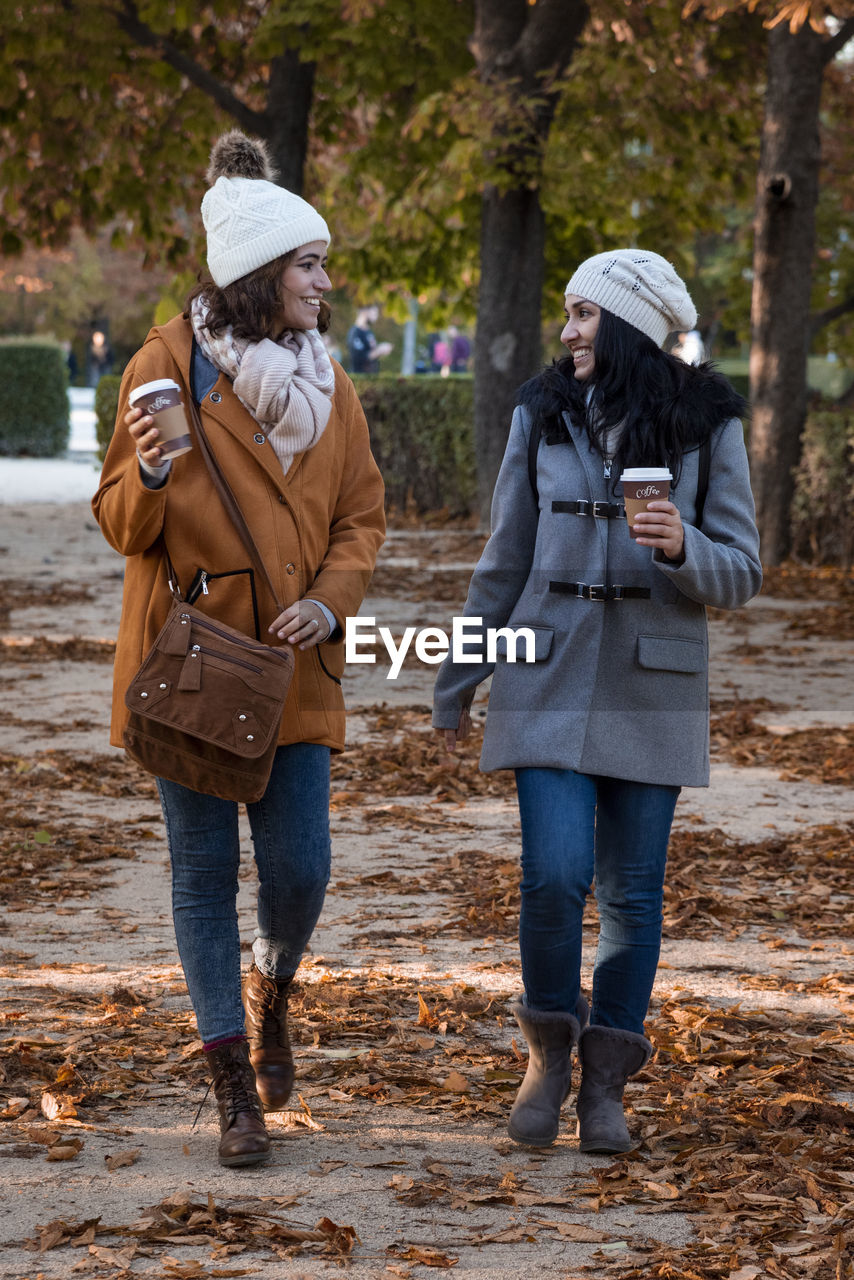 Best friends chatting in the park in the fall with their coffee to go. they're wearing a casual coat and a wool hat.