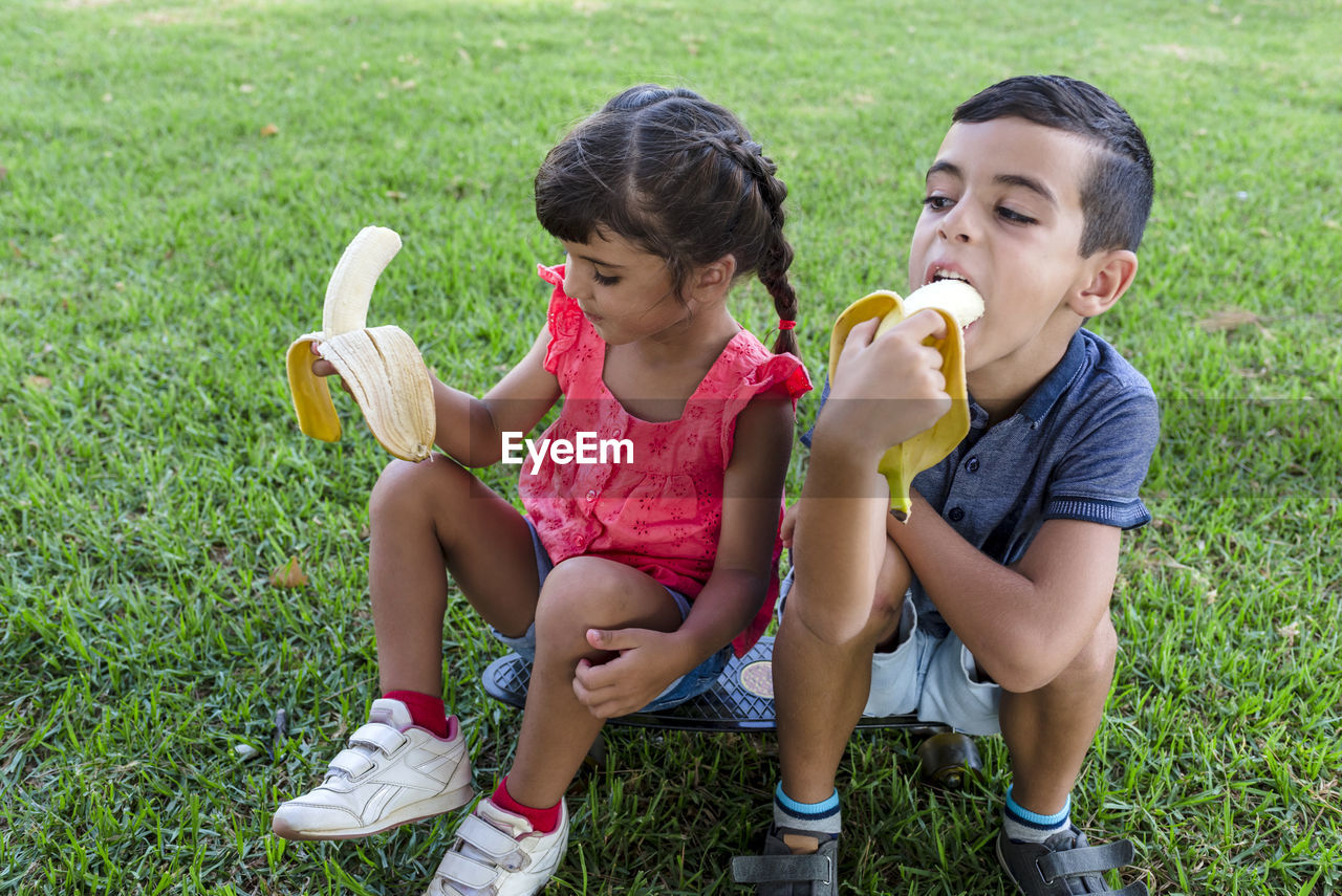 Two kids eating banana sitting outdoors on the grass in a park.