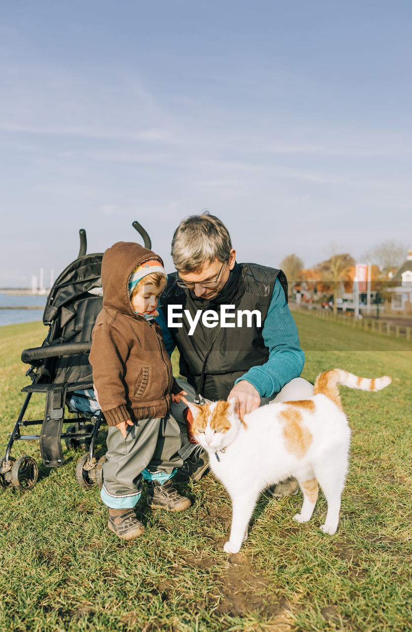 Grandfather with granddaughter looking at cat on grassy field against sky in park