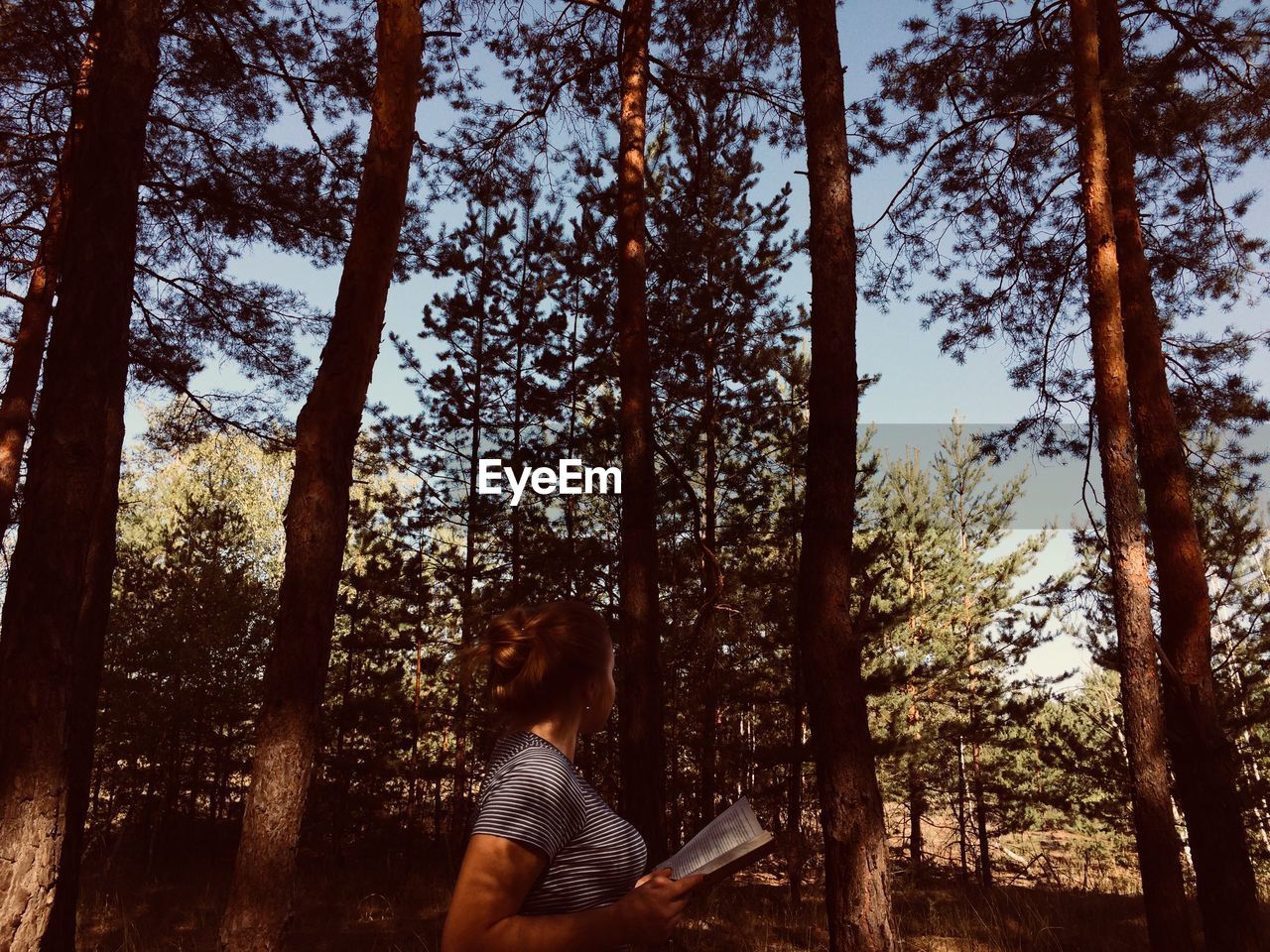 Woman holding book while standing against trees in forest