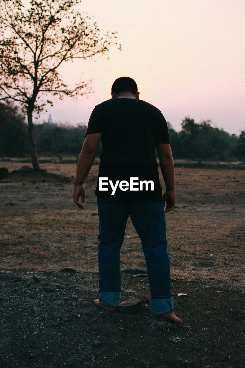 REAR VIEW OF MAN STANDING ON FIELD AGAINST SUNSET SKY