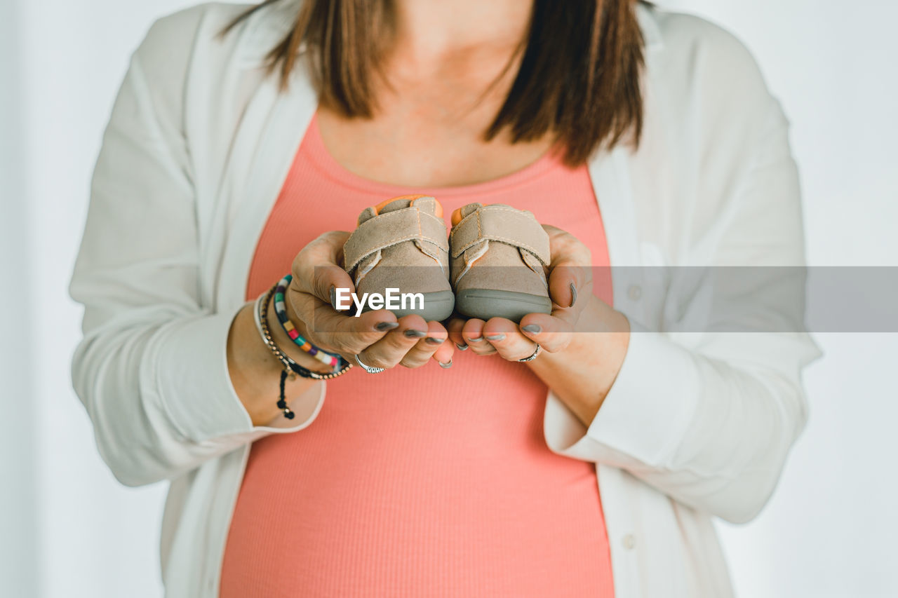 Midsection of pregnant woman holding baby shoes against white background