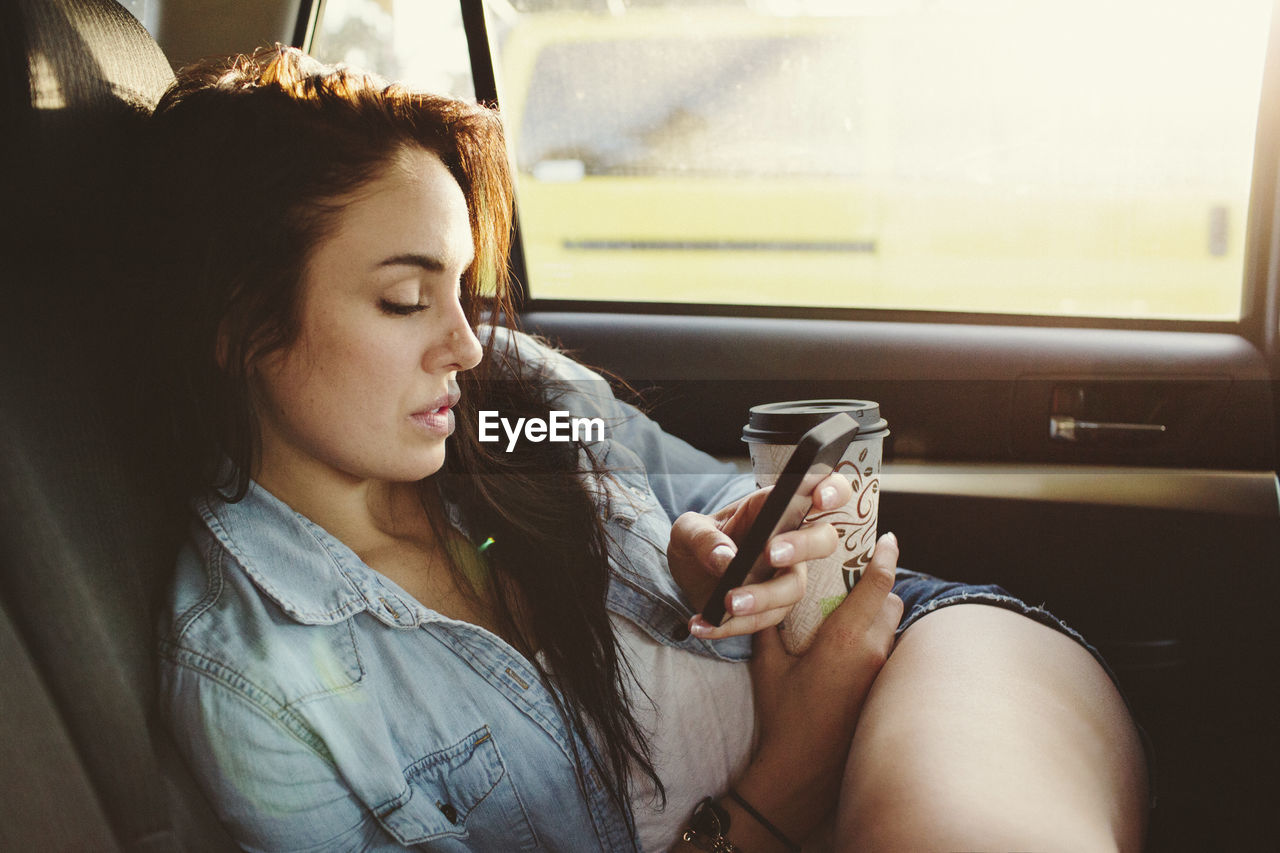 Woman with disposable cup using mobile phone while traveling in car