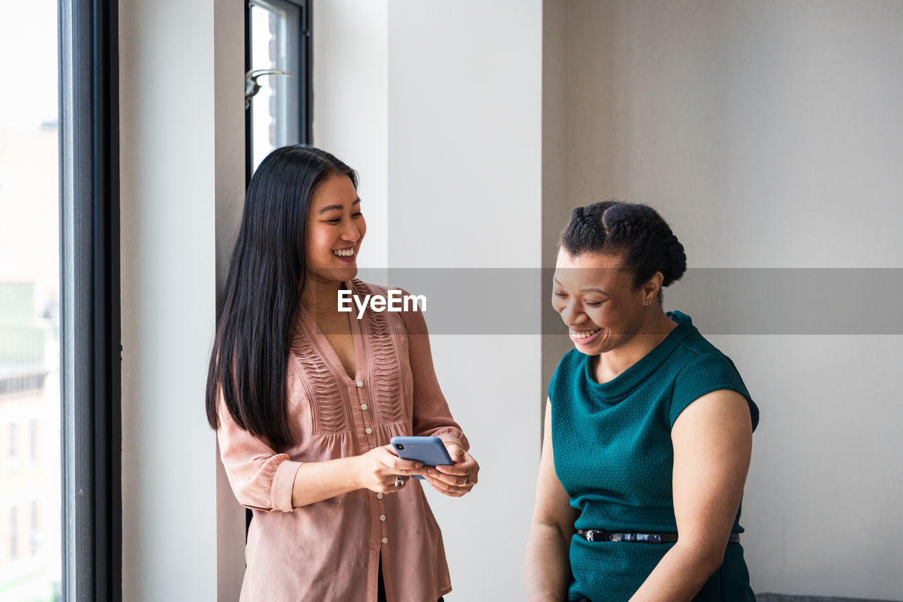 Smiling businesswoman showing mobile phone to female colleague by window at office