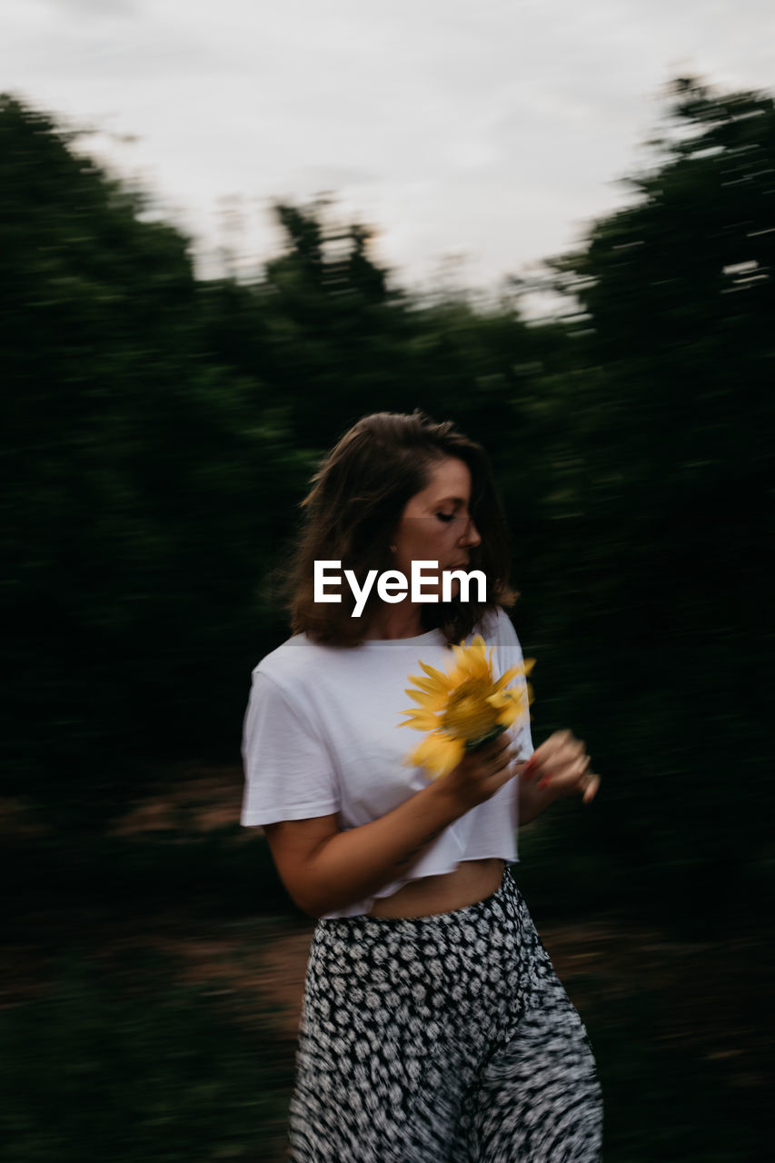 Side view of brown haired woman in light clothing with standing with flowers in hands amid green trees during summer vacation looking at camera