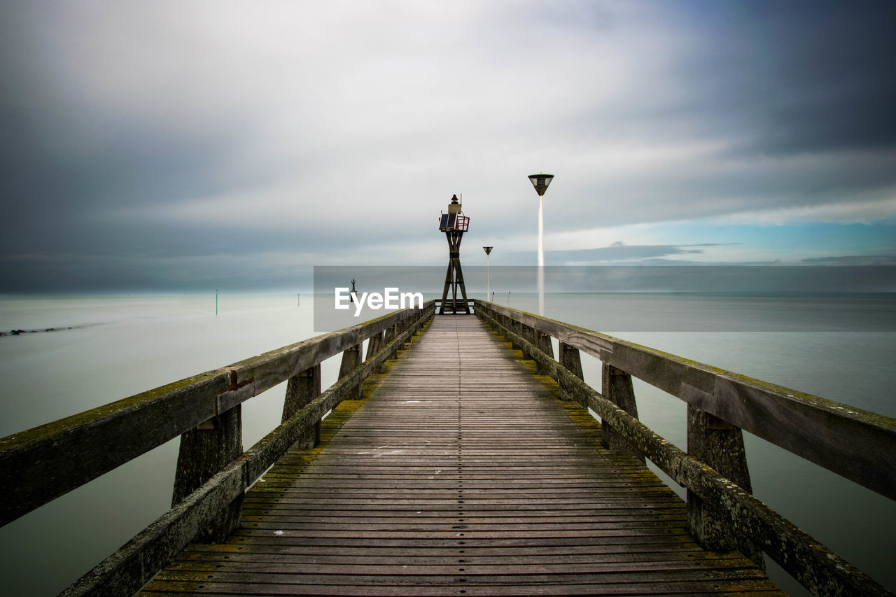 Empty jetty leading to sea against cloudy sky