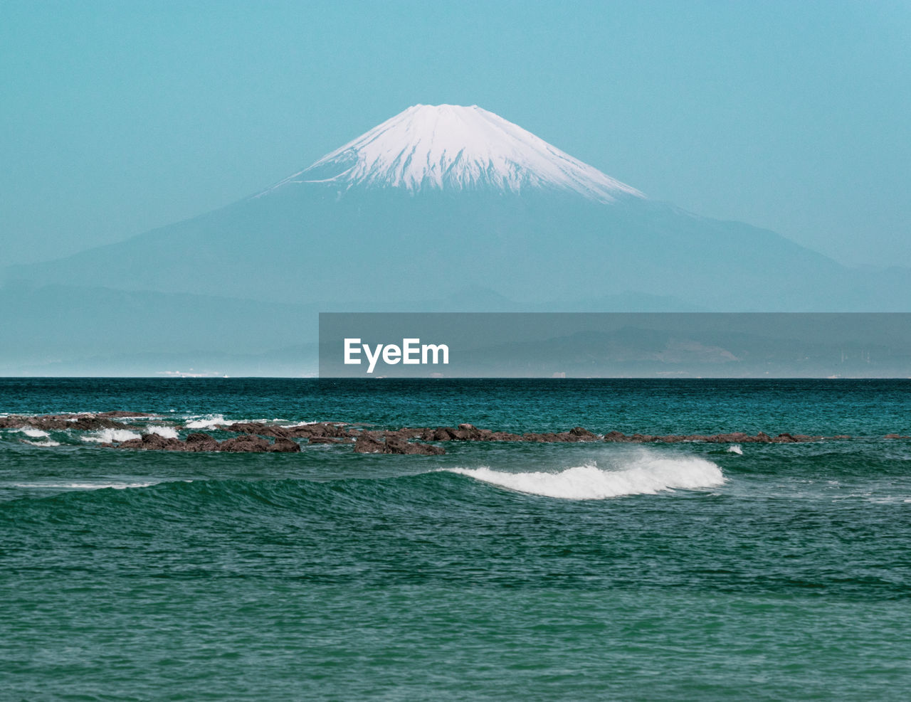 Scenic view of mt. fuji against hazy summer sky and ocean with waves in foreground.