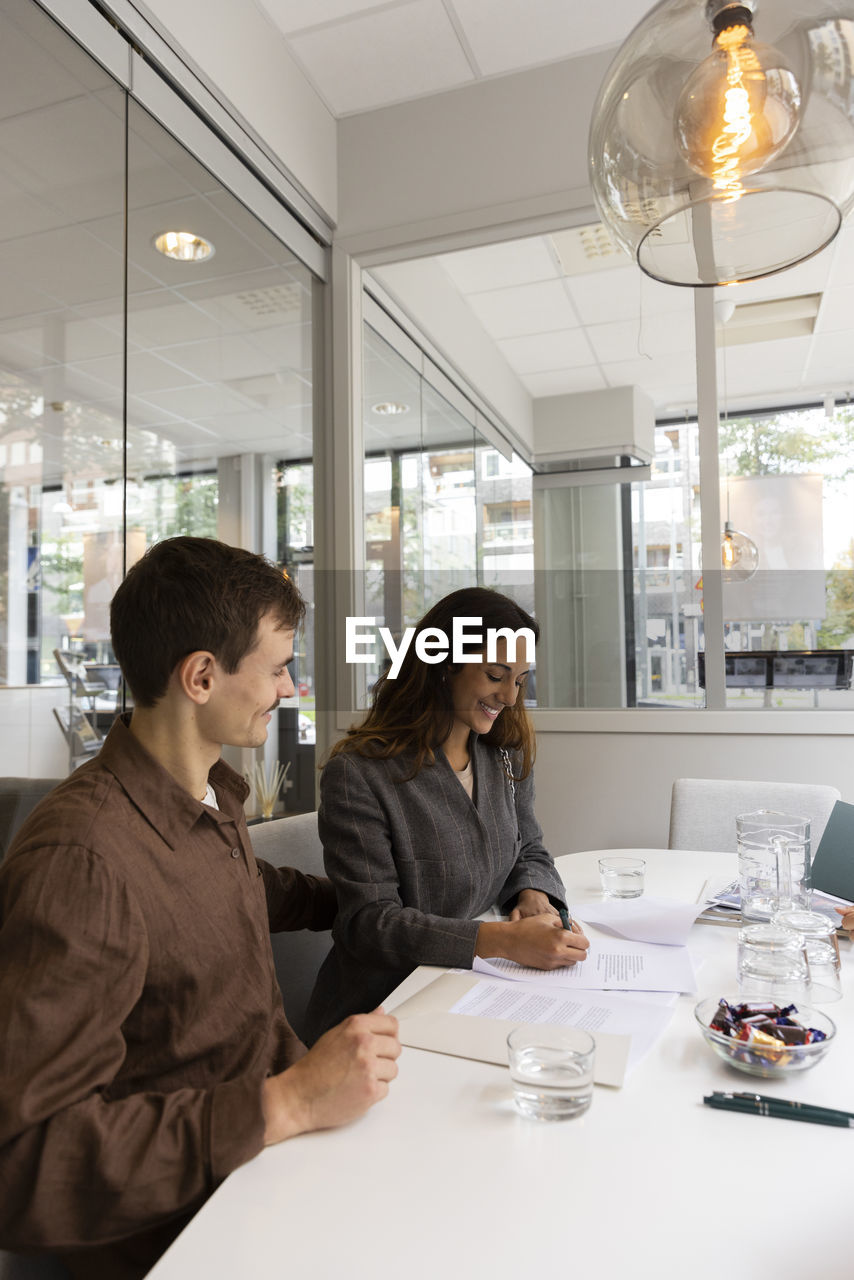 Smiling man looking at woman signing agreement at desk in real estate office