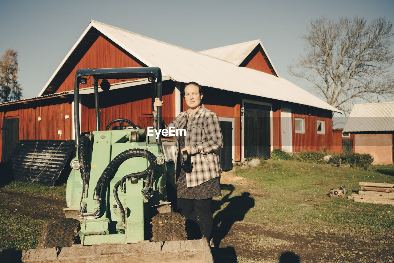 Portrait of mid adult female farmer standing by agricultural vehicle against barn