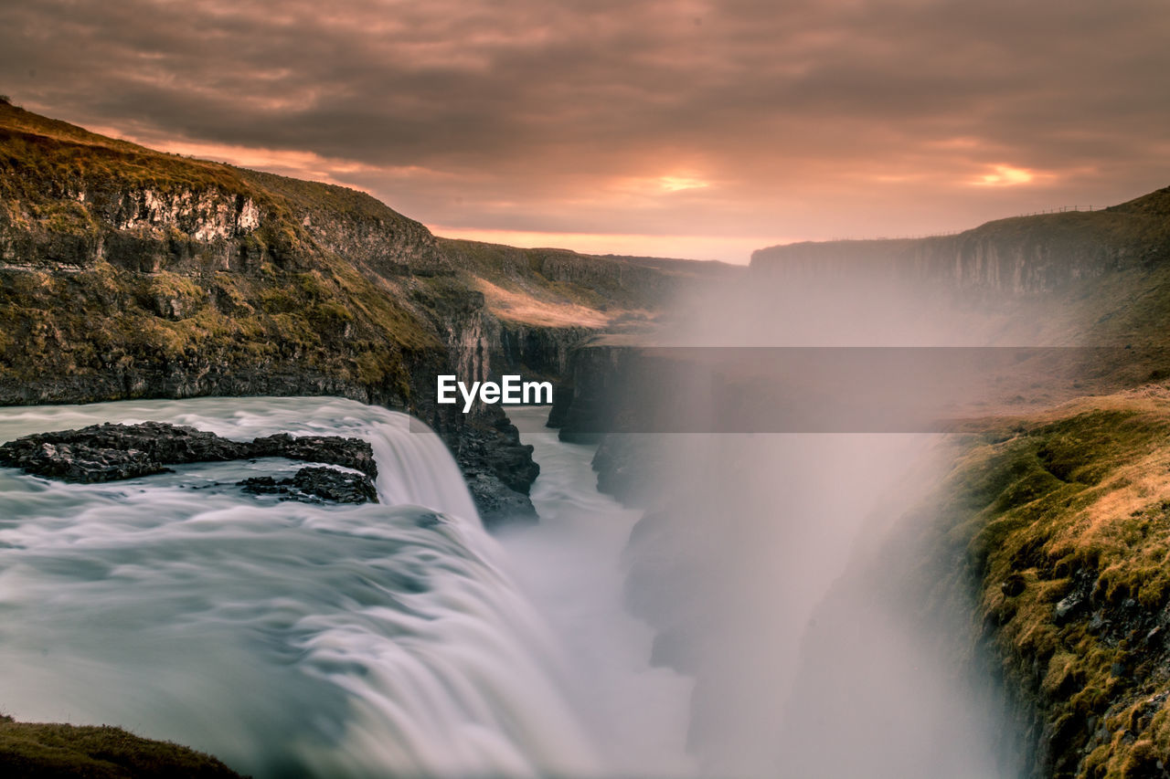 Scenic view of waterfall against cloudy sky during sunset