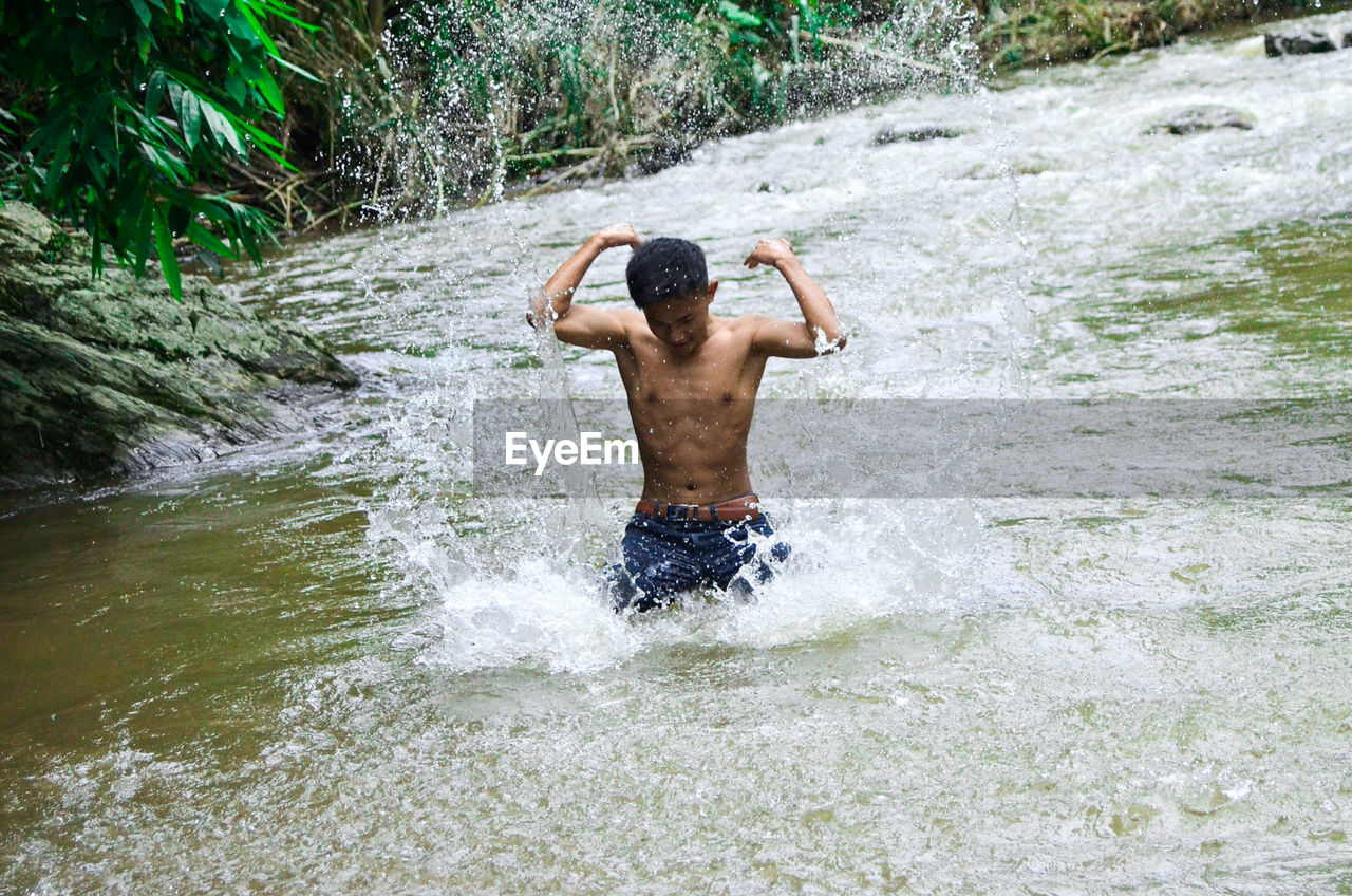 REAR VIEW OF SHIRTLESS MAN SPLASHING WATER IN WATERFALL