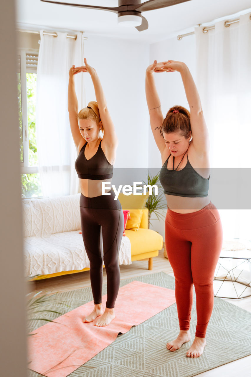 Tranquil females in sportswear standing in room and stretching bodies with raised arms during yoga class