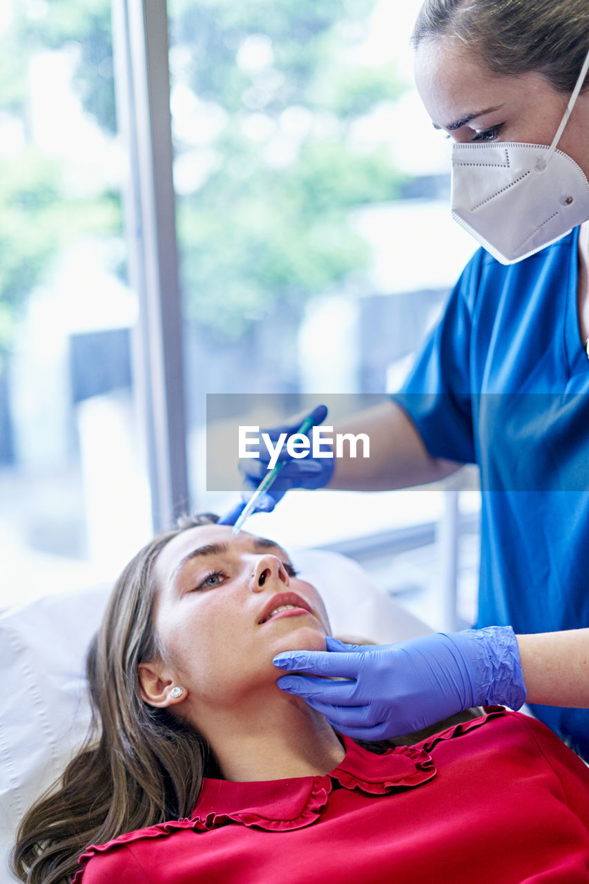 Esthetician in uniform and face mask injecting collagen into forehead of female client near window in office of beauty clinic.