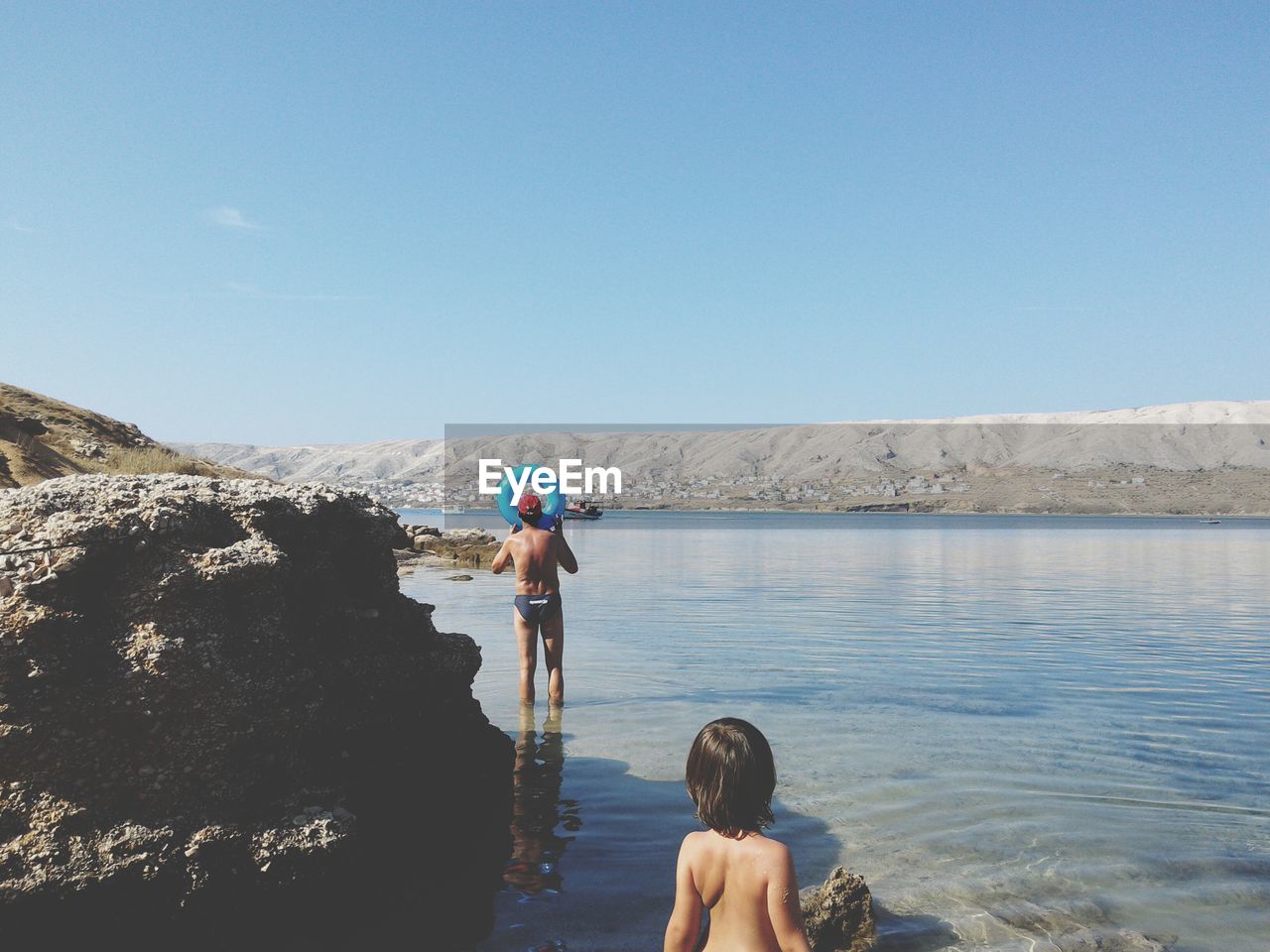 Shirtless father and son standing at riverbank against clear blue sky