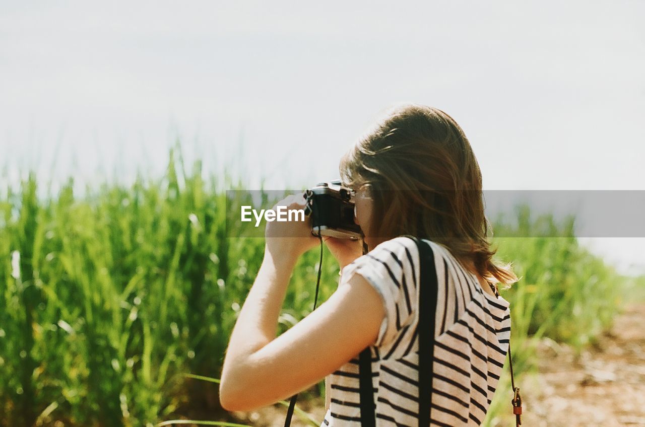 Side view of young woman photographing field against sky