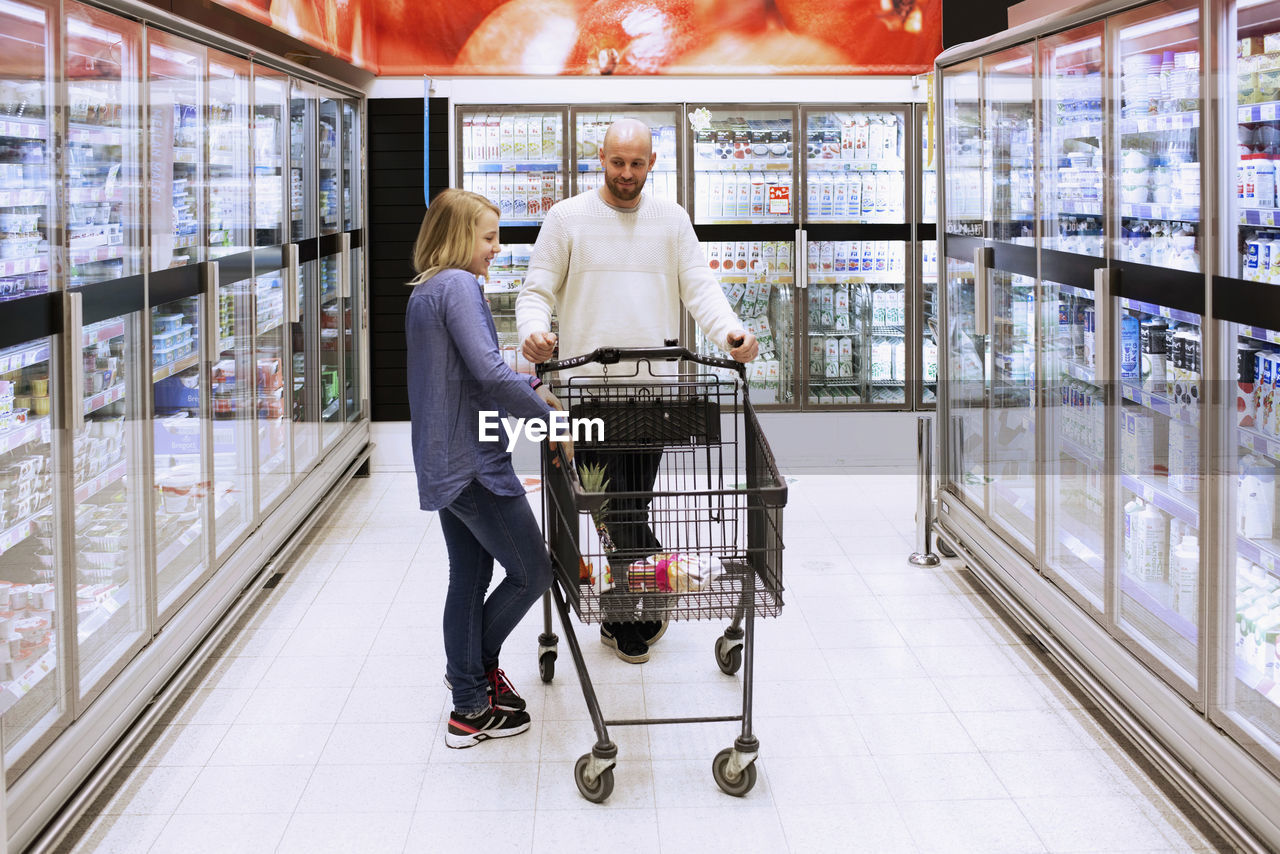 Full length of father and daughter with shopping cart in supermarket aisle