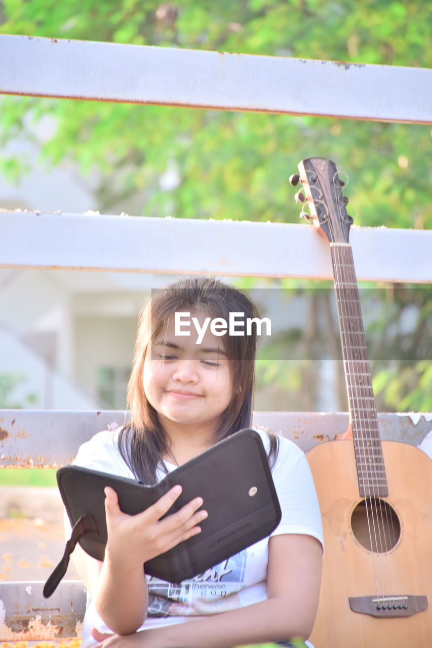 Smiling young woman using digital tablet by guitar against fence