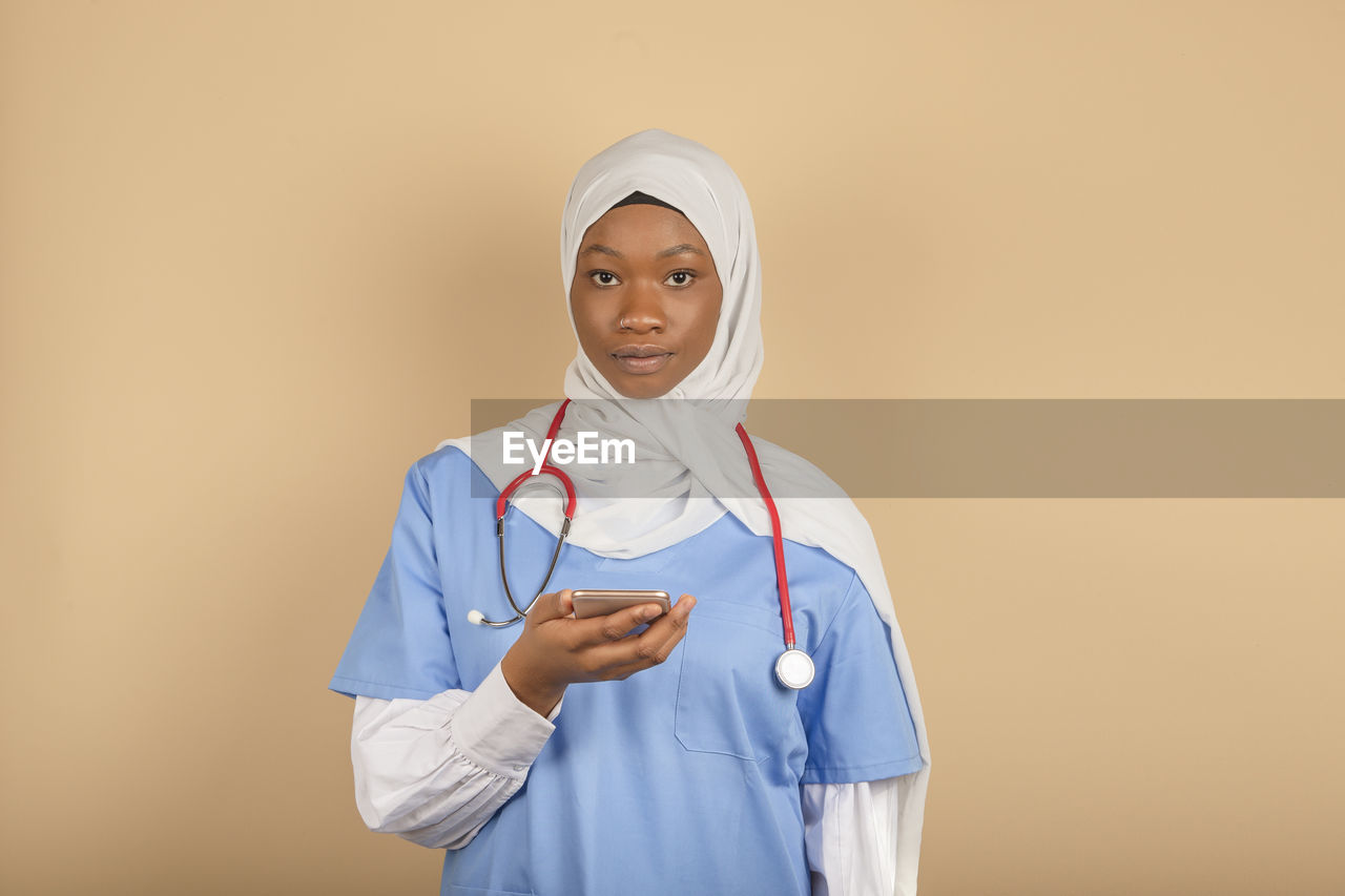 Young muslim nurse with mobile phone looking at camera on color background person