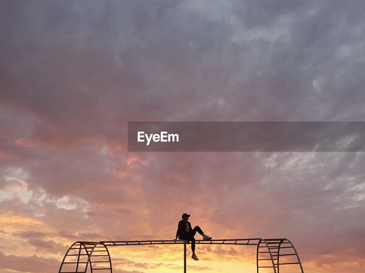 Silhouette man sitting on play equipment against sky during sunset