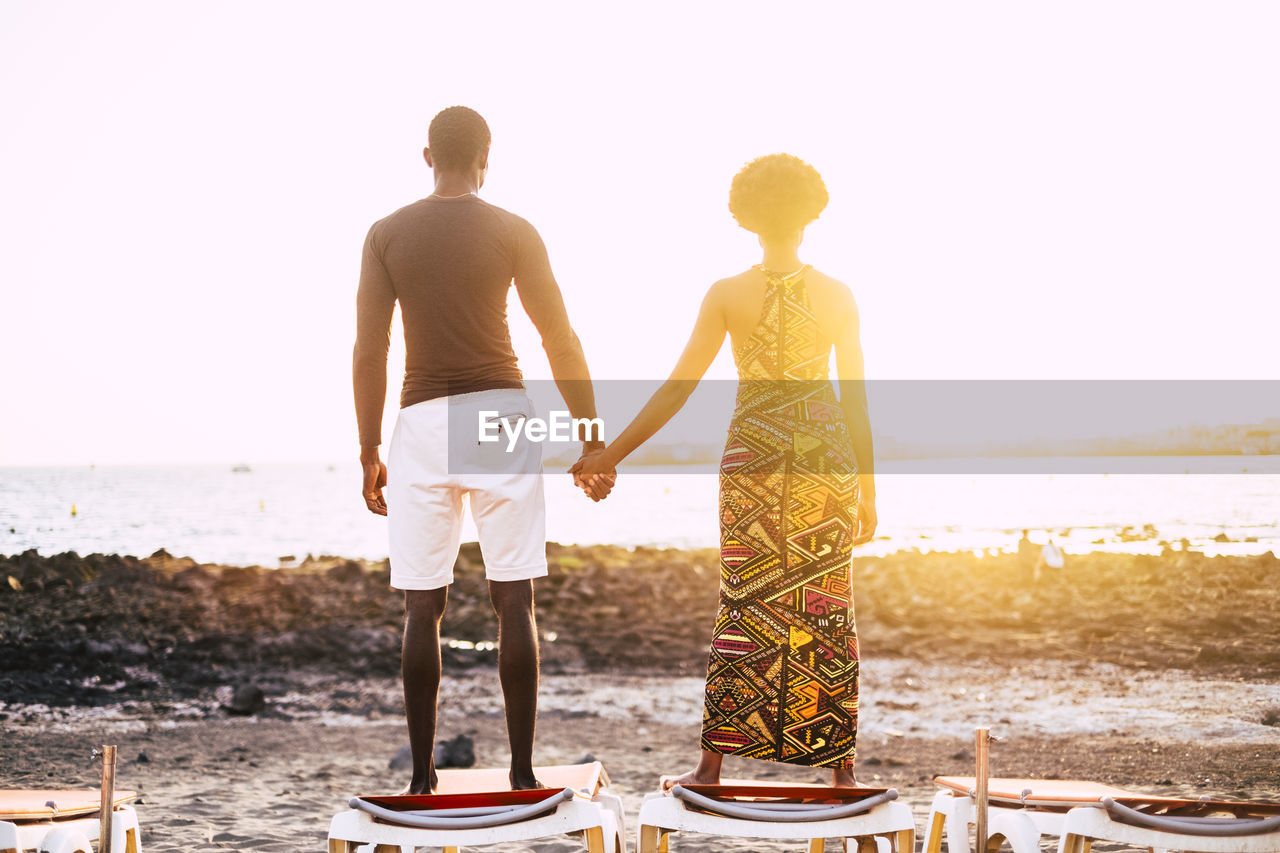 Rear view of couple holding hands while standing at beach against clear sky