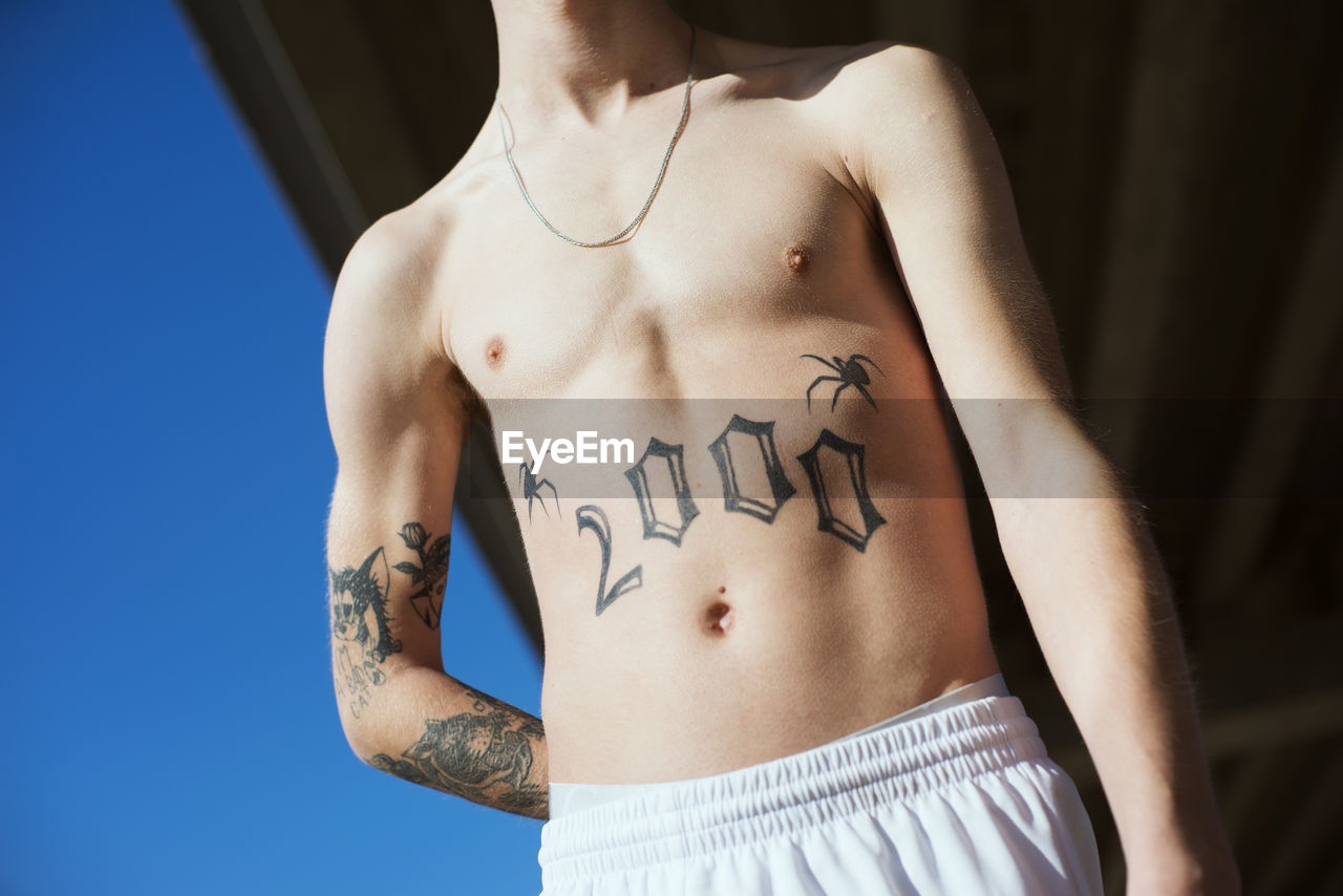 Low angle view of shirtless young man with tattoo standing outdoors