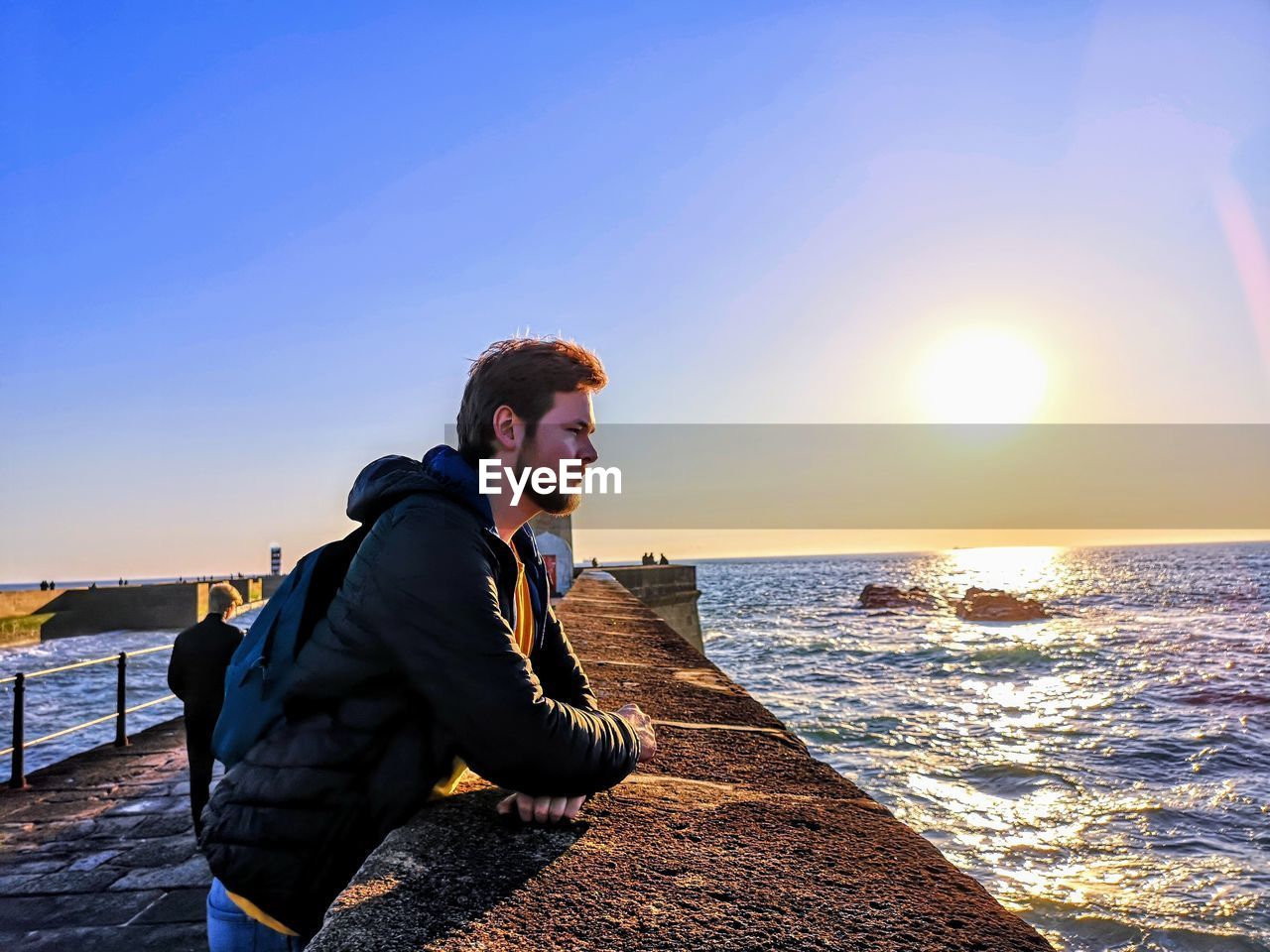 Young man leaning on retaining wall at beach against sky during sunset