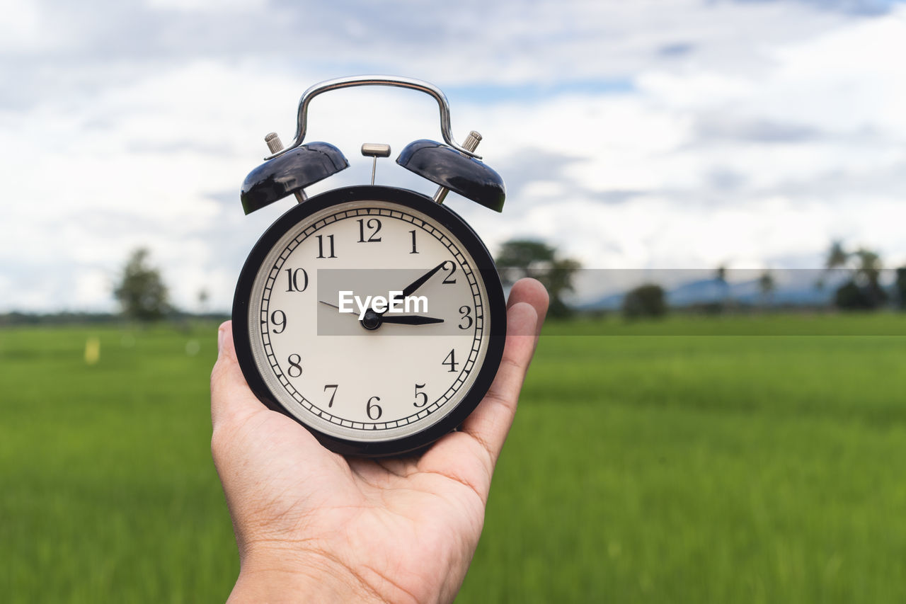 Close-up of hand holding alarm clock against sky