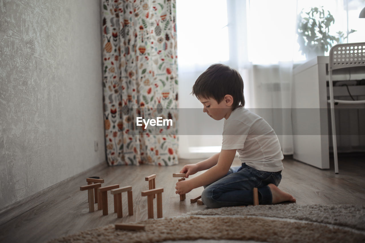 Boy caucasian brunette sad and serious plays on the floor, builds towers from wooden designer, 