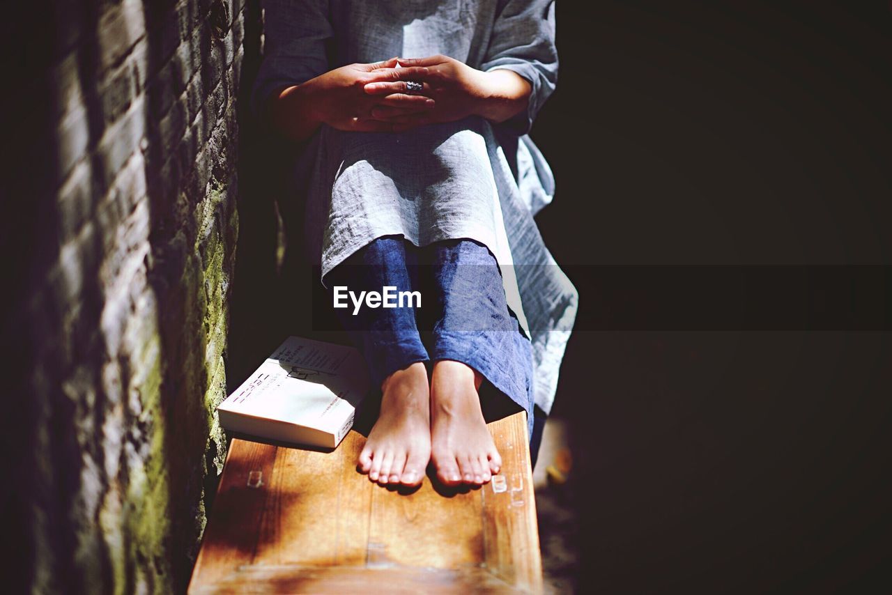 Low section of woman sitting with book on bench by wall